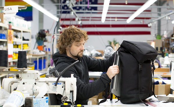 Nik sitting at a sewing machine table in the TOM BIHN factory with the American and Rainbow flags on the wall in the background. He is pulling a 15" Dell laptop out of the laptop compartment of the Shadow Guide 33.